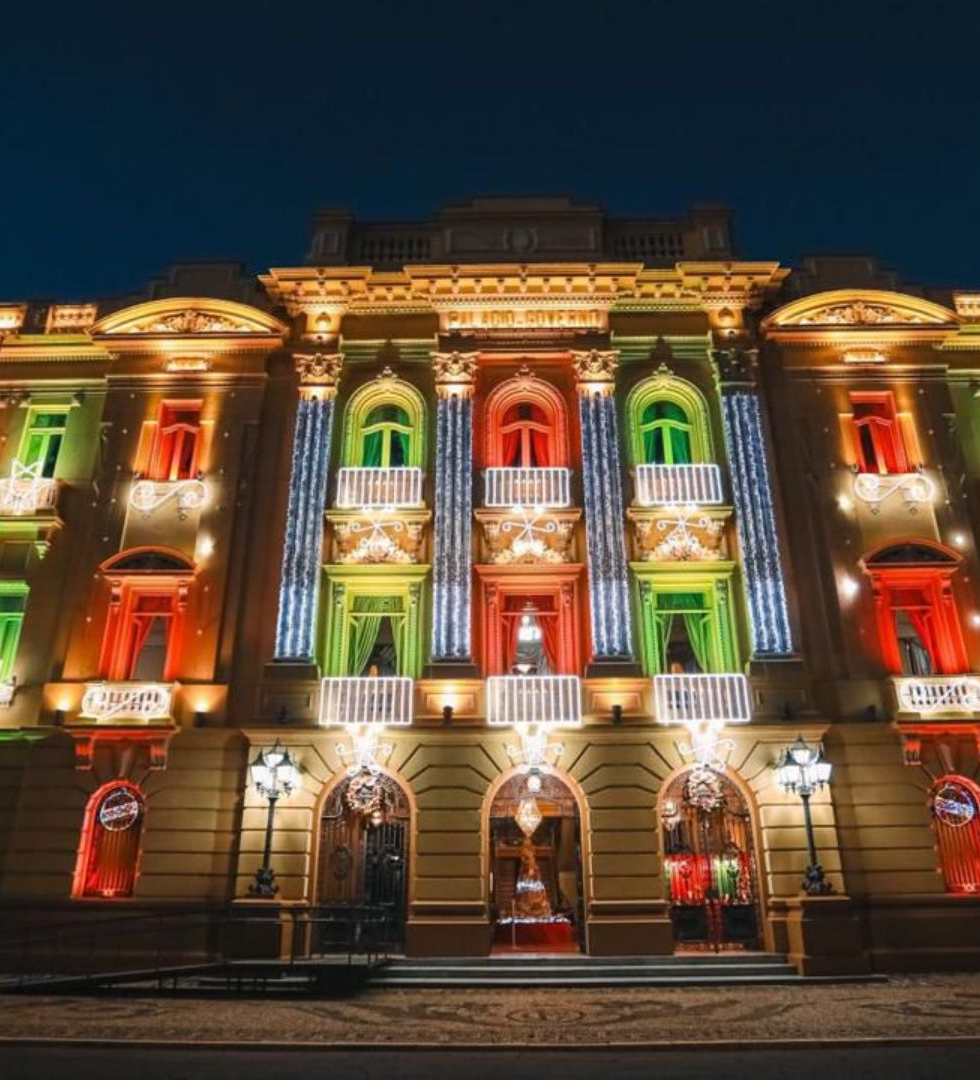 Fachada de prédio histórico à noite, iluminada em verde, vermelho e dourado, com palmeiras e céu escuro ao fundo.