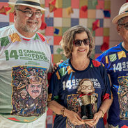 Três idosos sorridentes com chapéus e óculos, em camisas da 14ª Caminhada do Forró, posam diante de fundo colorido xadrez.