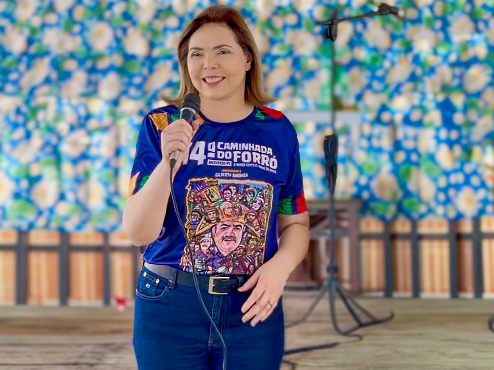 Mulher sorridente fala ao microfone em palco colorido, usando camiseta azul com texto 14ª Caminhada do Forró.