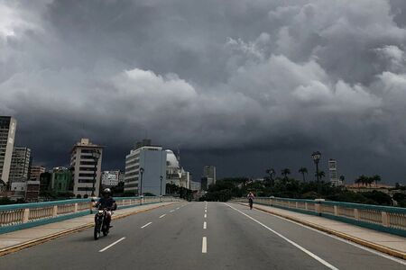 Céu nublado sobre ponte em cidade, com motociclista e pedestre. Prédios no fundo, atmosfera de tempestade iminente.