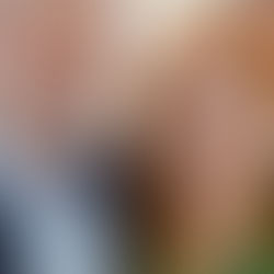 Dois adultos sorriem em um ambiente interno. Um homem de barba branca veste terno azul e uma mulher de cabelo ruivo usa blusa verde.