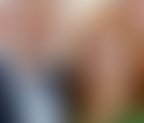 Dois adultos sorriem em um ambiente interno. Um homem de barba branca veste terno azul e uma mulher de cabelo ruivo usa blusa verde.