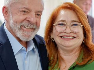 Dois adultos sorriem em um ambiente interno. Um homem de barba branca veste terno azul e uma mulher de cabelo ruivo usa blusa verde.