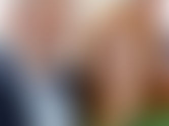 Dois adultos sorriem em um ambiente interno. Um homem de barba branca veste terno azul e uma mulher de cabelo ruivo usa blusa verde.