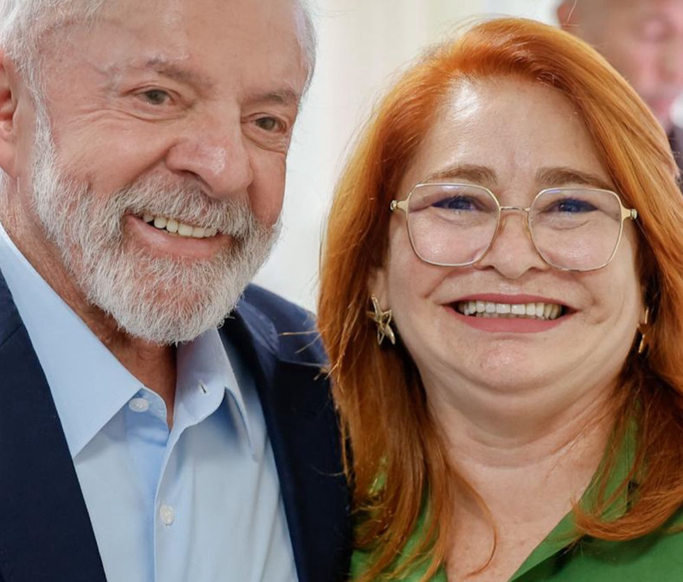 Dois adultos sorriem em um ambiente interno. Um homem de barba branca veste terno azul e uma mulher de cabelo ruivo usa blusa verde.