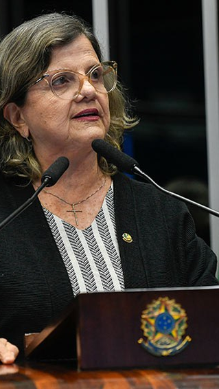 Mulher fala em pódio no Senado, com microfones e bandeira do Brasil ao fundo. Ela usa óculos e veste roupa escura. Cena formal.