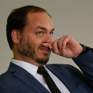 Homem de terno azul toca o nariz com a mão, em pose pensativa, diante de fundo neutro.