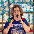 Mulher de óculos fala ao microfone, com camiseta colorida escrita Caminha do Forró, diante de fundo azul floral.