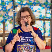 Mulher de óculos fala ao microfone, com camiseta colorida escrita Caminha do Forró, diante de fundo azul floral.