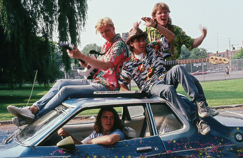 A group of 1980s teens sit on top of a paint splattered car holding instruments.