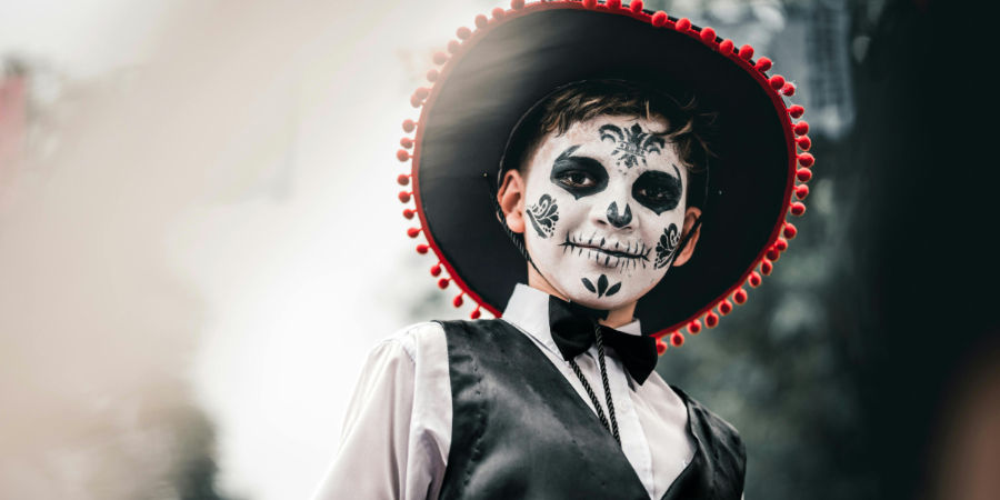 Child in calavera skull face paint wearing a black sombrero with red pom-poms, vest, and bow tie ready for Dia de los Muertos activities