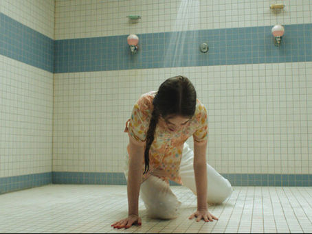 A girl in a floral top and white pants kneels on a tiled shower-room floor as water runs from a single showerhead.