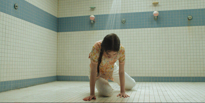 A girl in a floral top and white pants kneels on a tiled shower-room floor as water runs from a single showerhead.