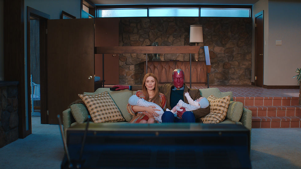 Couple sits on a green couch in a stone-walled living room, each holding a baby, with a red-faced man in a brown jacket beside them.