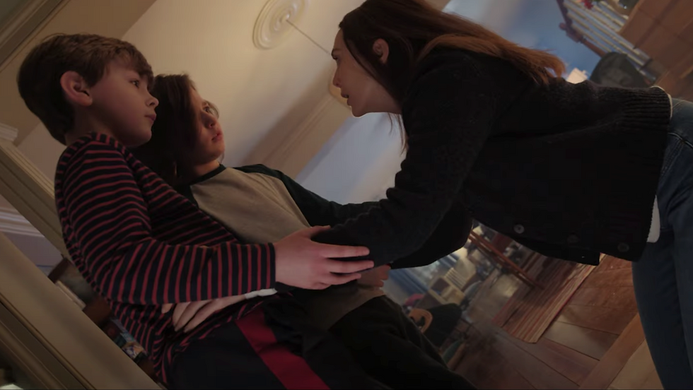 Woman leans toward two seated children in a hallway, holding one boy's arm, tense but caring.