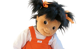 Demenzpuppe Emelie mit schwarzen Zöpfchen und enem orangen Latzkleid und einem weißen T-Shirt