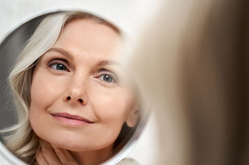 Happy 50s middle aged woman model touching face skin looking in mirror reflection. Smiling