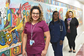3 diverse female staff members, in scrubs and professional attire standing in front of a huge colorful mural in one of the building's hallways.