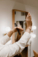 A hairstylist at work in a salon setting. The stylist, wearing a white blazer and a gold bracelet, is cutting hair with professional scissors in one hand while holding a comb and a section of blonde/light brown hair in the other. The scene is captured in warm, soft lighting with blurred circular light elements in the background, creating an elegant, professional atmosphere. A wooden-framed mirror is visible in the background, reflecting the salon space. The composition captures the precision and artistry involved in professional hairstyling at TRU Hair Salon in Colorado Springs.