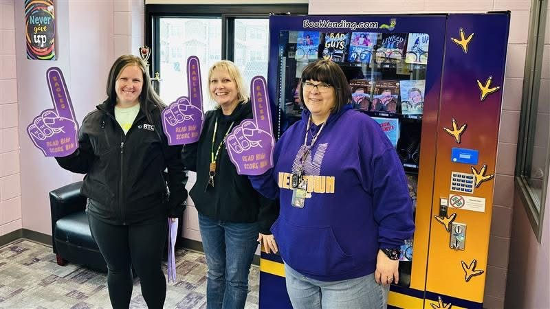 Women wearing foam fingers stand next to a book vending machine.