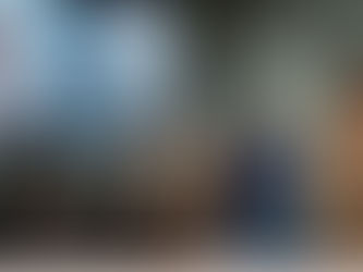 A medium-angle, full shot captured by artificial intelligence shows a high-end fashion show set within a stark, industrial space with concrete walls and exposed metal trussing. On a large LED screen at the left, two virtual, digital women with smooth features and bob haircuts are dressed in modern, structured attire—one wearing a flared tan leather dress and the other in a glistening silver fringed dress. Below the screen, a row of diverse, casually-dressed guests are seated, observing as four male models with long hair walk toward the viewer. The models are casually dressed in neutral beige jackets and jeans, with one in a striking blue denim jacket and sweater. At the far right, the back of a black-haired guest in a tan ribbed dress stands, adding depth to the scene. Soft natural daylight from large windows illuminates the event.