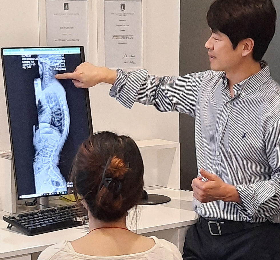 A Chiropractor points to a spine X-ray on a monitor while explaining to a seated woman in an office. Certificates hang on the wall.