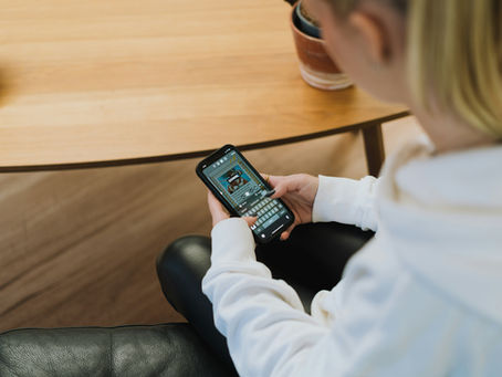 Person in a white hoodie using a smartphone, typing a message for a Story. Seated on a black leather chair near a wooden table. Relaxed setting.
