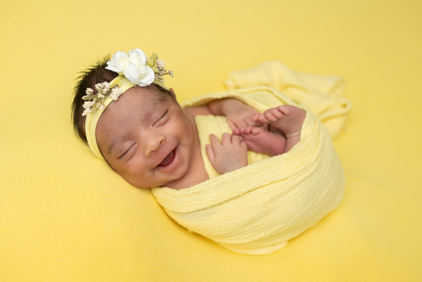A beautiful newborn is posed delicately for a photo, wrapped snugly in a soft yellow wrap that complements their tiny frame. The gentle fabric creates a warm and cozy cocoon around the baby, enhancing their natural innocence. A matching yellow floral headband adorns their head, adding a sweet touch to the serene look. The baby's eyes are closed peacefully, and a hint of a smile graces their lips, exuding tranquility. The background is softly blurred, allowing the focus to remain on the baby, capturing the pure essence of new life and the joy that comes with it. The overall composition radiates warmth and tenderness, creating a heartwarming image.