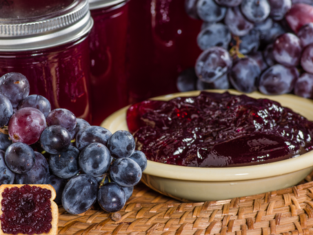 Grapes and jars of jam on a woven mat. A bowl of grape jelly and a slice of bread with jam. Rich purple hues dominate the scene.