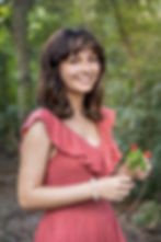 High School Senior in a pink dress smiling, holding small red flowers in a lush, green outdoor setting in Houston, TX. She's wearing a pearl bracelet.