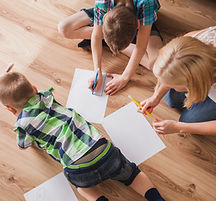 Zwei Kinder und eine Erwachsene sitzen am Boden und schreiben etwas auf weiße Blätter, Ansicht aus der Vogelperspektive