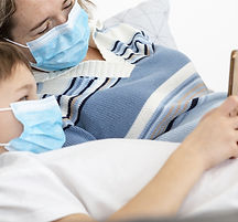 Kind und Erwachsene liegen in Bett und lesen gemeinsam ein Buch, beide tragen Mundschutz