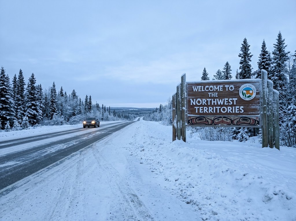 An outdoor, winter photograph of the rustic wooden welcome sign at the border of the Northwest Territories, Canada, on the side of a snow-covered highway. The large wooden sign reads "WELCOME TO THE NORTHWEST TERRITORIES" and features traditional Indigenous-style artwork and territorial branding, partially frosted with snow. The surrounding landscape is heavily blanketed in snow, with evergreens lining the roadside under an overcast, subarctic sky. A single SUV is visible driving in the distance on the plowed, wet asphalt road, capturing the vast and icy northern environment.