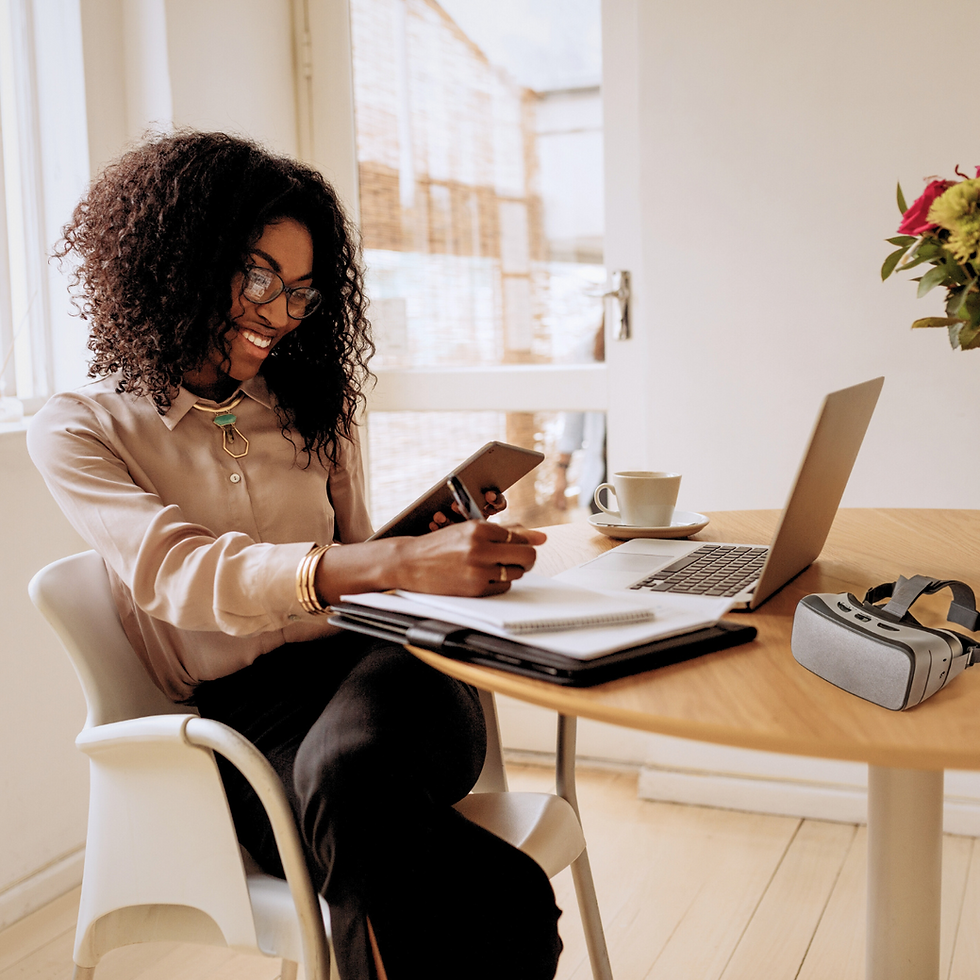 Creative, professional diverse woman with black curly hair wearing a modern and fashionable outfit while writing in on a notebook in front of her laptop and VR headset.