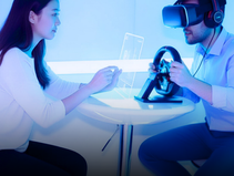 A woman and a man with VR headset at a table, man holding a steering wheel. Futuristic setting with blue lighting and digital screens.