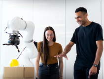 Two business people smile at a robotic arm placing a yellow ball on a box. They're in a bright room, creating an atmosphere of excitement.