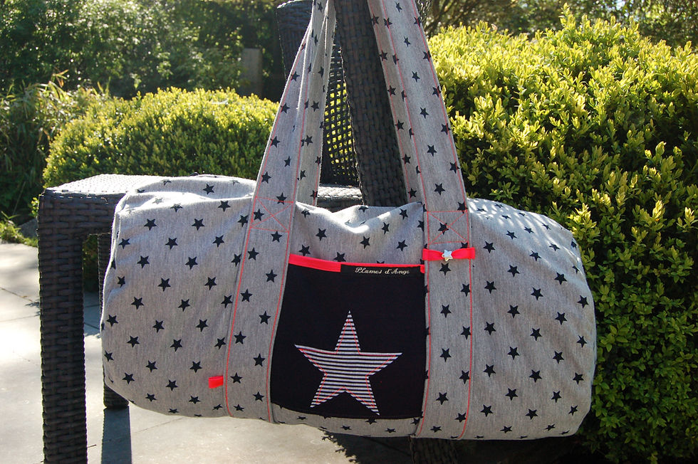 Sac gris avec étoiles noires, poche avant étoilée, texte Plumes d'Ange. Plantes vertes et chaise en osier en arrière-plan. Ambiance décontractée.