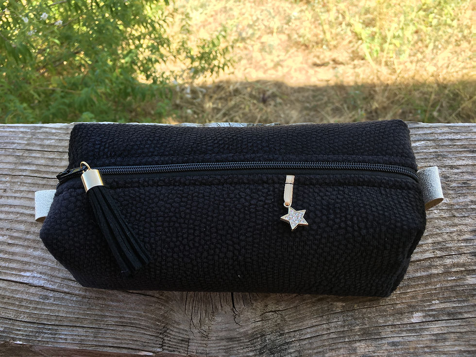 Pochette noire avec breloque étoile et pompon, posée sur une table en bois. Arrière-plan de verdure floue et lumière naturelle.