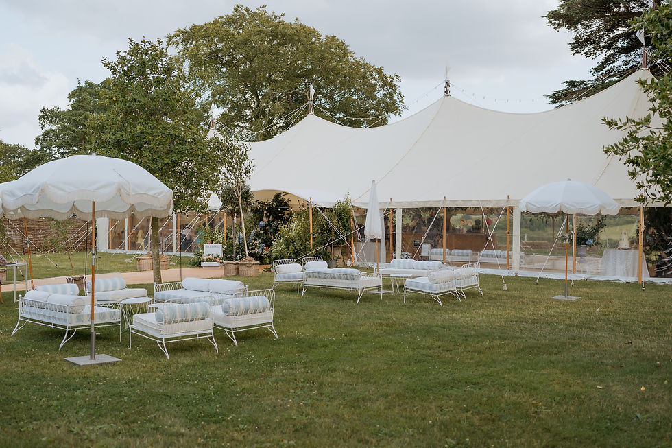 White marquee in a manicured lawn with white furniture and parasols. Trees in background. A "Welcome" sign visible. Calm, outdoor setting.
