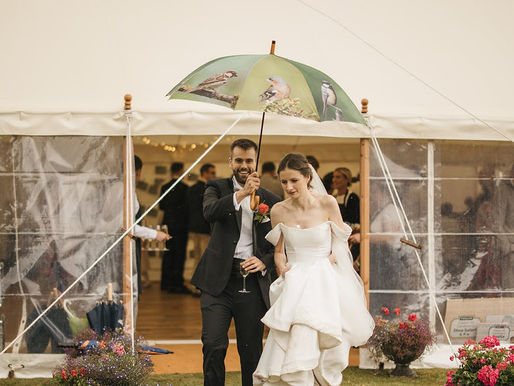 Bride and groom walk on grass under a bird-patterned umbrella, smiling. They're exiting a marquee with fairy lights, surrounded by flowers.