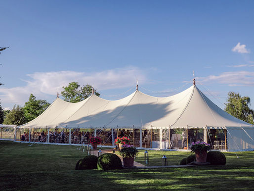 Large white event marquee on a sunny day, surrounded by trees and grass. People are visible inside. Potted plants and lanterns adorn the path.