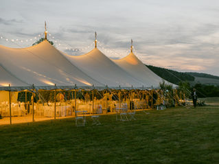 Outdoor wedding under a large illuminated marquee, with decorated tables and guests mingling. Twilight sky and hills in the background.