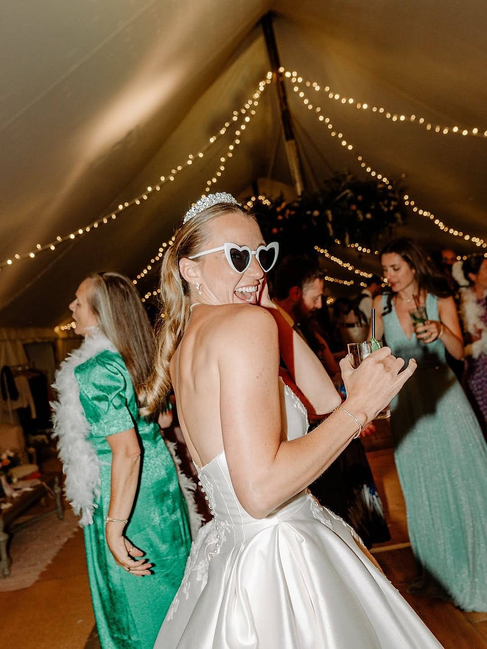 Bride in a white dress, heart sunglasses, and tiara smiling and holding a drink in a pole and canvas wedding marquee with fairy lights in the background.