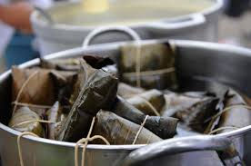 Steamed banana leaf-wrapped parcels fill a metal pot, likely tamales or similar, in a kitchen setting. The background is blurred.