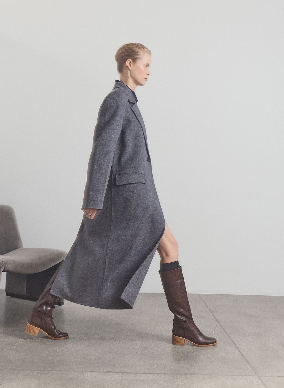 Blond woman walking in front of a grey velvet chair wearing a grey coat and cognac knee-high boots