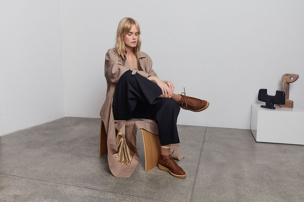 Blond woman seated looking at her suede shoe in a gallery with sculptures on a plinth