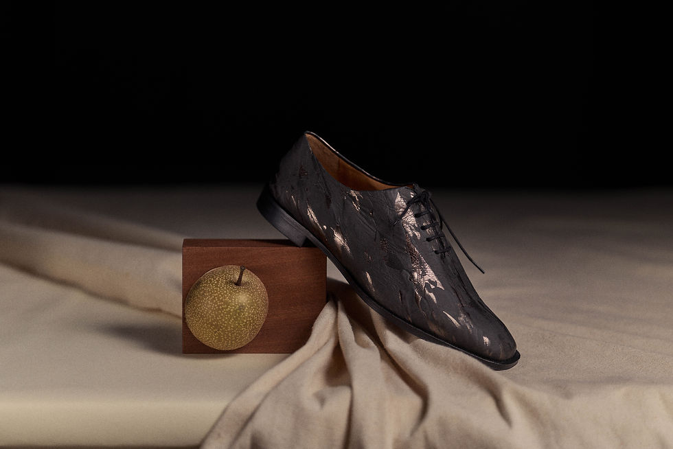 Still-life photo of an oxford shoe supported by a wooden cube, with an Asian pear inside, on a table with wavy canvas fabric.