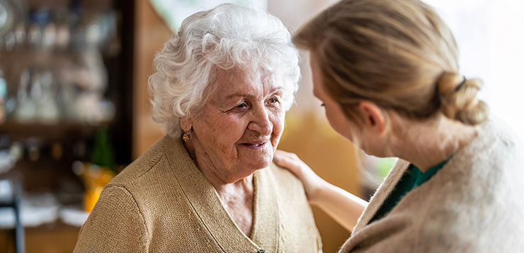 Older woman smiles as a younger woman comforts her indoors, hand on shoulder, warm and tender mood.