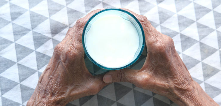 Elderly hands holding a cup of milk over a gray and white geometric patterned tablecloth. Tranquil and homely atmosphere in Aged Care