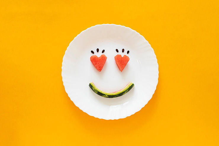 White plate on bright orange background with a playful face made from fruit slices, smiling like a happy emoji